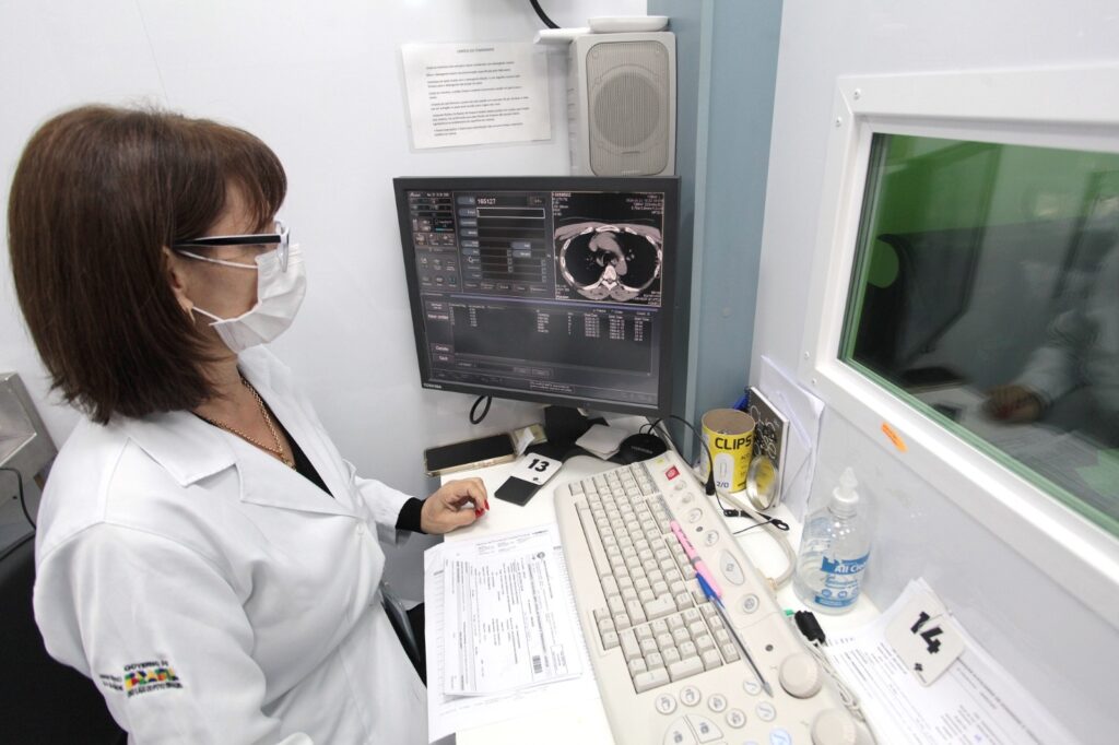 A imagem mostra uma técnica de radiologia, vestindo jaleco branco e máscara facial, sentada em uma estação de trabalho em um ambiente clínico. Ela está olhando para um monitor que exibe uma imagem de tomografia computadorizada (TC) do tórax humano. Na mesa à sua frente, há um teclado, alguns controles específicos para o equipamento, um celular, um frasco de álcool em gel e uma caneca com a palavra "CLIPS". Ao lado, há uma grande janela com vidro verde, pela qual a mão de outra pessoa é visível. Na parede, há um cartaz com instruções detalhadas sobre a limpeza do tomógrafo, escrito em português, orientando sobre o uso de detergente neutro e cuidados para não danificar o equipamento. O ambiente é limpo, organizado e bem iluminado, transmitindo um aspecto profissional e técnico.