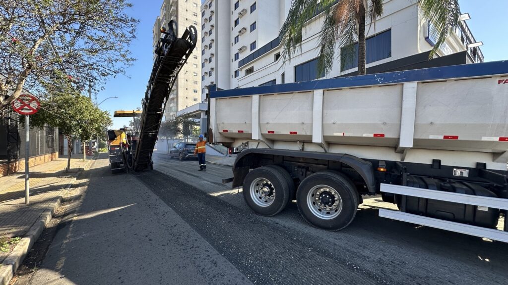 A imagem mostra uma cena de pavimentação ou manutenção de estrada. No centro, há uma máquina fresadora de asfalto em operação, retirando a camada superior da via. A máquina está conectada a uma esteira transportadora que direciona o material fresado para a caçamba de um caminhão branco, posicionado à direita da imagem. Um trabalhador, vestido com colete de segurança laranja e capacete branco, está parado entre a fresadora e o caminhão, aparentando supervisionar o processo. Ao fundo, há um edifício moderno de vários andares com janelas, indicando que a cena ocorre em uma área urbana. Também é possível ver uma árvore de palmeira, sugerindo um clima tropical ou subtropical. Um sinal de proibição de estacionamento está fixado em um poste à esquerda, próximo ao meio-fio. O céu está claro e azul, com luz solar forte que cria sombras bem definidas no chão e nos veículos. A imagem transmite uma sensação de trabalho em andamento, com a poeira e detritos levantados pela fresadora destacando a atividade pesada na rua.