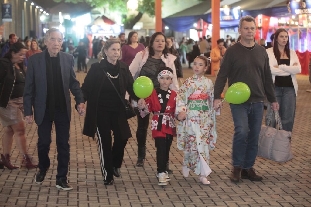A imagem mostra um grupo de pessoas caminhando juntas em um ambiente ao ar livre durante a noite, provavelmente em um festival ou evento cultural. O grupo é composto por seis pessoas de diferentes idades, sugerindo uma família ou um grupo próximo. Entre elas, há duas crianças vestindo roupas tradicionais japonesas: o menino usa um happi vermelho com detalhes brancos e uma faixa na cabeça, enquanto a menina veste um quimono com estampa floral. As crianças seguram balões verdes. Os adultos estão vestidos com roupas casuais e de clima ameno, como casacos e suéteres. Ao fundo, é possível ver barracas iluminadas, outras pessoas e elementos típicos de uma feira ou festival. A atmosfera parece alegre e movimentada, com luzes artificiais que iluminam o local durante a noite. No canto superior direito, há uma placa parcialmente visível com as palavras "COREIA DO SU", indicando que o evento pode ter alguma relação com a cultura coreana ou ser um festival multicultural.