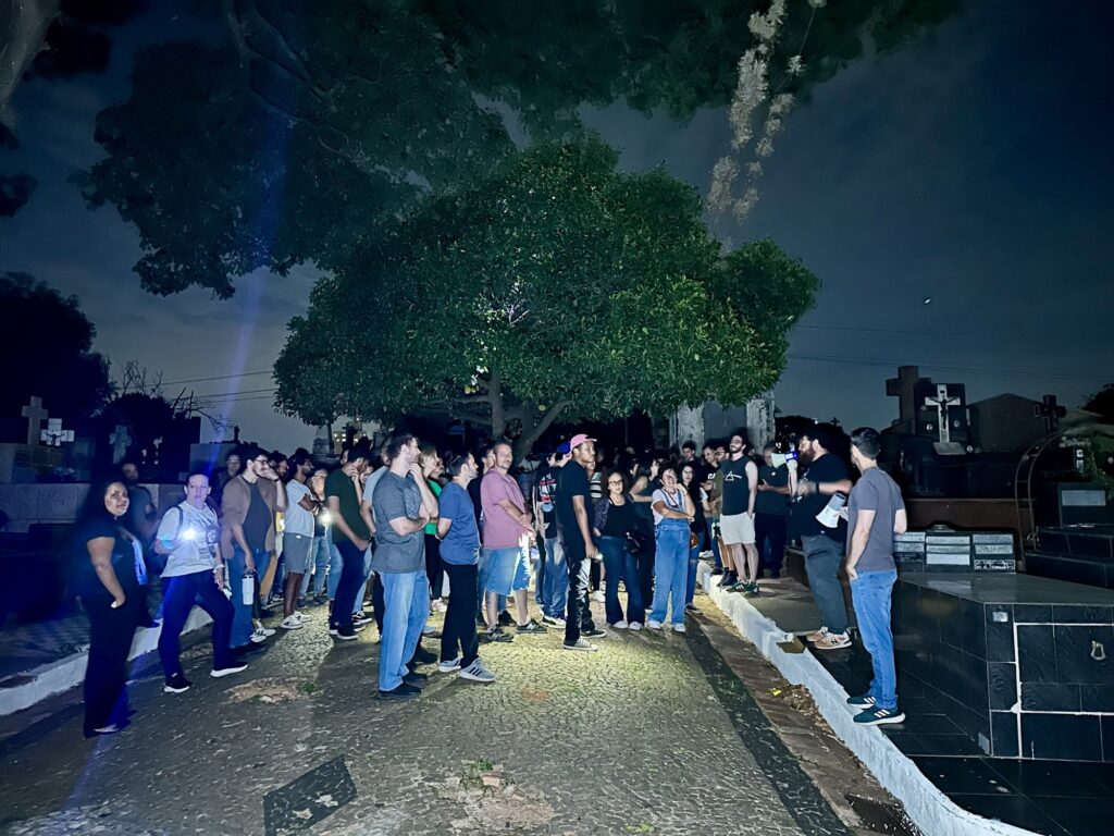 A imagem mostra um grupo de pessoas reunidas em um cemitério durante a noite. Elas parecem estar participando de uma visita guiada ou evento organizado, pois estão atentas e algumas seguram lanternas que iluminam o caminho e o ambiente ao redor. O cenário inclui um caminho de pedras no primeiro plano e várias lápides e cruzes ao fundo, típicas de um cemitério. A iluminação é artificial, com as lanternas criando áreas de luz e sombra, conferindo uma atmosfera dramática e um pouco misteriosa. As cores predominantes são escuras, com tons de azul e cinza do céu noturno e os tons sombrios do cemitério. As pessoas estão vestidas de forma casual, indicando que não se trata de um evento formal. Ao fundo, há árvores com folhagem densa que contribuem para o clima do local. A perspectiva da foto é levemente elevada, mostrando o grupo e o caminho de cima para baixo. A cena sugere que se trata de uma visita noturna ao cemitério, possivelmente um passeio histórico ou turístico.