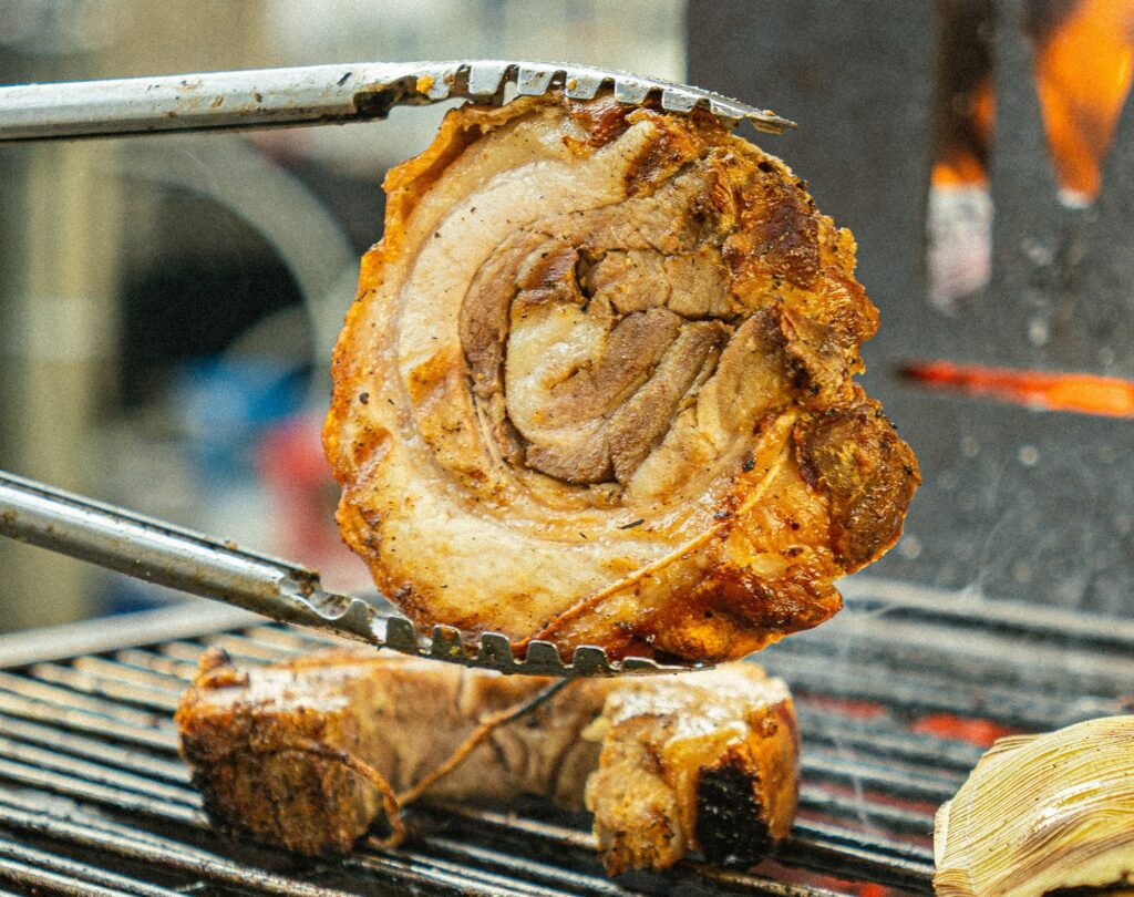 A imagem mostra um close de uma peça de barriga de porco assada sendo segurada por um pegador de metal sobre uma grelha. A barriga de porco está enrolada, com a pele dourada e crocante, e camadas visíveis de carne e gordura. O pegador tem dentes que ajudam a segurar firmemente a carne. A grelha está posicionada acima de brasas incandescentes, indicando que o alimento está sendo preparado em fogo direto. O fundo está desfocado, sugerindo um ambiente de cozinha ao ar livre ou uma churrasqueira. A iluminação quente valoriza a textura suculenta e apetitosamente dourada da carne. A imagem transmite a sensação de um prato saboroso e recém-preparado na brasa.