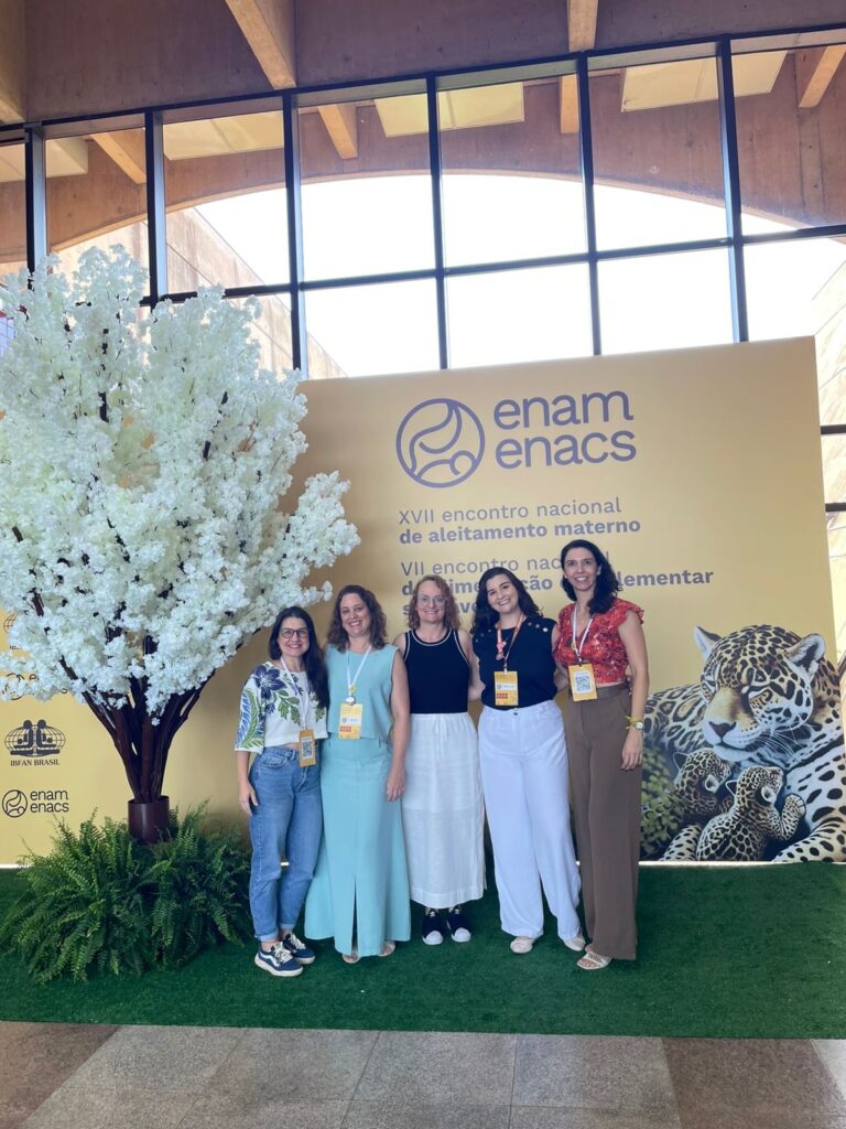 A imagem mostra cinco mulheres posando para uma foto em um ambiente interno, provavelmente em um evento. Elas estão em pé, alinhadas em frente a um grande banner amarelo, que contém o texto "XVII encontro nacional de aleitamento materno" e os logos "enam" e "enacs". Também há a imagem de uma onça com dois filhotes no banner. À esquerda da imagem, há uma árvore artificial com flores brancas e algumas samambaias verdes ao redor. O espaço parece ser amplo, com janelas grandes que deixam entrar muita luz natural, além de uma estrutura de madeira visível no teto. As mulheres estão sorrindo e usando roupas casuais, algumas com crachás pendurados no pescoço, indicando que são participantes ou organizadoras do evento. A atmosfera é alegre e acolhedora, sugerindo um encontro relacionado à promoção do aleitamento materno.
