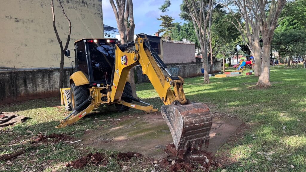 A imagem mostra uma retroescavadeira amarela da marca Caterpillar (CAT) em um ambiente ao ar livre, possivelmente um parque ou área de lazer. A máquina está posicionada sobre uma superfície gramada, com os estabilizadores abaixados para dar suporte e o braço da retroescavadeira apoiado em uma pequena laje de concreto. Ao fundo, é possível ver um parquinho colorido com brinquedos, algumas árvores e um muro de concreto desgastado. A retroescavadeira é o foco principal da imagem, destacando-se pelo seu tamanho, cor vibrante e detalhes da estrutura metálica. A cena está iluminada pela luz natural do dia, criando sombras que ressaltam as formas da máquina e do ambiente ao redor.