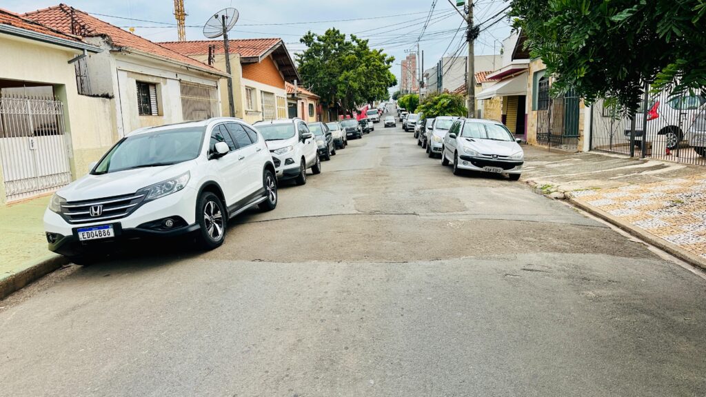 A imagem mostra uma rua residencial estreita, provavelmente em um bairro tranquilo. As casas são pequenas, de um andar, com telhados de telhas vermelhas e paredes claras. Na calçada, há vários carros estacionados dos dois lados da rua, incluindo um Honda branco e um Peugeot prata. A rua é asfaltada, mas apresenta marcas e desgastes visíveis. O céu está nublado, sugerindo um dia cinzento. Fios elétricos cruzam a imagem na parte superior, mostrando a infraestrutura típica das áreas urbanas. A cena está vazia de pessoas, transmitindo uma sensação de calma e silêncio.