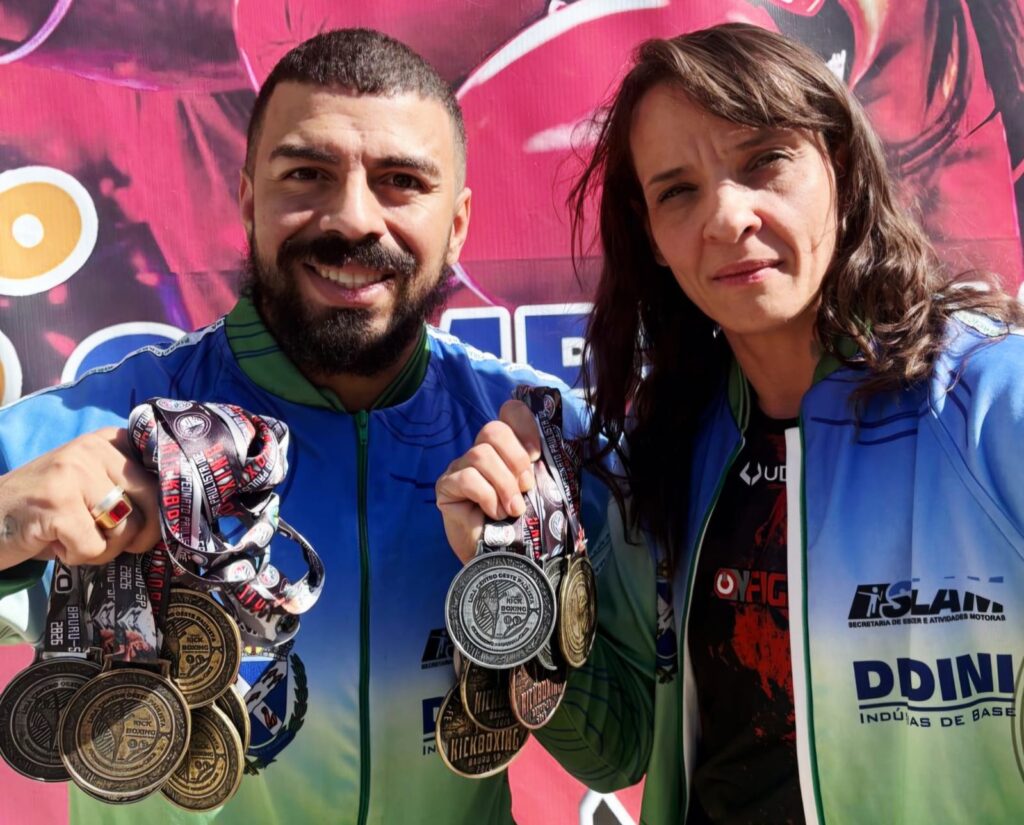 A imagem mostra um homem e uma mulher posando juntos, ambos exibindo várias medalhas penduradas no pescoço e segurando algumas na mão. Eles estão vestidos com jaquetas esportivas nas cores azul e verde, com logos e inscrições relacionadas a competições esportivas. O homem tem barba e está sorrindo, enquanto a mulher tem uma expressão mais séria. Ao fundo, há um banner rosa com elementos circulares amarelos e brancos, sugerindo um ambiente de evento ou premiação. As medalhas indicam participação e conquistas em campeonatos de artes marciais, como kickboxing e boxe, com inscrições que mencionam "Campeonato Paulista", "Bauru-SP" e outras localidades. A imagem transmite uma sensação de orgulho e realização esportiva.