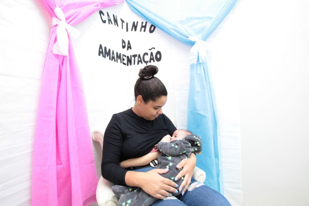 A imagem mostra uma cena acolhedora em um ambiente interno, onde uma mãe está amamentando seu bebê. Ela está sentada em uma poltrona clara, confortável, vestindo uma blusa preta de mangas longas e jeans rasgados. O bebê está enrolado em um macacão cinza com capuz e pequenos detalhes coloridos, repousando no colo da mãe. Ao fundo, a parede branca é decorada com duas cortinas de tecido, uma rosa à esquerda e outra azul clara à direita, que criam um contraste suave e agradável. No centro da parede, entre as cortinas, está escrito em letras maiúsculas e escuras "CANTINHO DA AMAMENTAÇÃO", indicando que o local é um espaço dedicado para amamentação. A iluminação é clara e uniforme, transmitindo uma sensação de calma e conforto. A imagem enfatiza o cuidado e a intimidade do momento entre mãe e filho, em um ambiente pensado para oferecer privacidade e tranquilidade.