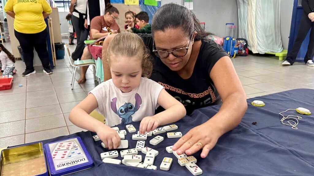Esta imagem captura um momento educativo e de interação em um ambiente que parece ser uma sala de aula ou um centro comunitário. * Em destaque, uma criança pequena, vestindo uma camiseta com a estampa do personagem Stitch, está concentrada jogando dominó sobre uma mesa coberta por uma toalha azul escura. * Ao lado dela, uma mulher adulta, usando óculos e uma camiseta preta, observa atentamente e auxilia a criança na organização das peças de dominó, criando um ambiente de apoio e aprendizado. * O cenário ao fundo mostra outras pessoas, incluindo crianças e adultos, em atividades diversas, sugerindo um espaço de convivência coletiva. Há também uma caixa de dominó visível no canto inferior esquerdo, indicando o material utilizado na atividade.