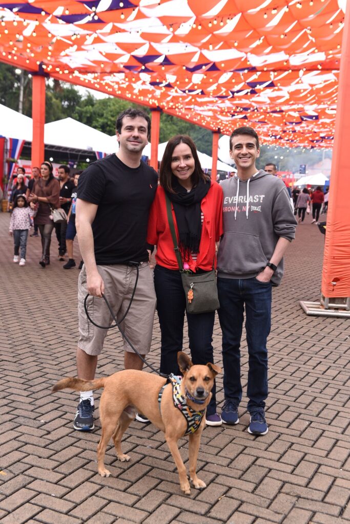 A imagem mostra três pessoas e um cachorro em um ambiente ao ar livre, possivelmente em um evento ou festival. Eles estão em um espaço pavimentado, com uma estrutura de cobertura feita de painéis laranja e brancos, decorada com luzes em cordão. À esquerda, um homem está segurando a guia de um cachorro de porte médio, cor caramelo, que usa um peitoral estampado. Ele veste camiseta preta, bermuda bege e tênis. No centro, uma mulher está sorrindo, vestindo uma blusa listrada, um casaco vermelho e uma echarpe preta, com uma bolsa pequena pendurada no ombro. À direita, outro homem veste um moletom cinza com a palavra "EVERLAST" estampada e jeans azuis. Ao fundo, é possível ver outras pessoas e barracas brancas, reforçando a ideia de que se trata de um evento público ou feira. A atmosfera parece descontraída e amigável.