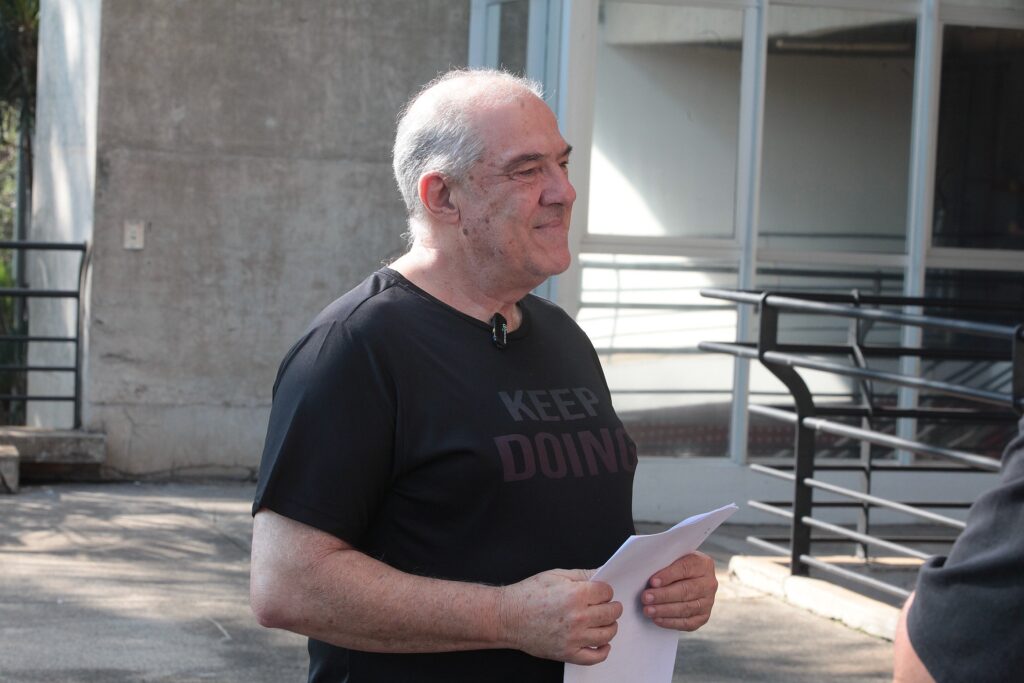 Esta imagem mostra um homem de meia-idade, com cabelos brancos curtos, em um ambiente externo durante o dia. Ele está de perfil, voltado para a direita, e apresenta uma expressão facial serena, com um leve sorriso. O homem veste uma camiseta preta com a frase "KEEP DOING" estampada no peito, em letras maiúsculas, e um pequeno microfone de lapela está preso à gola da camisa. Ele segura algumas folhas de papel branco com as duas mãos, sugerindo que pode estar se preparando para uma leitura, uma entrevista ou uma apresentação. O cenário ao fundo consiste em uma estrutura arquitetônica de concreto e metal, com uma grade preta e uma porta de vidro, indicando um ambiente possivelmente acadêmico ou corporativo. A iluminação é natural e lateral, destacando as feições do homem e projetando sombras suaves no local. No canto inferior direito, é possível ver parcialmente o braço e o ombro de outra pessoa, sugerindo a presença de um interlocutor ou cinegrafista.