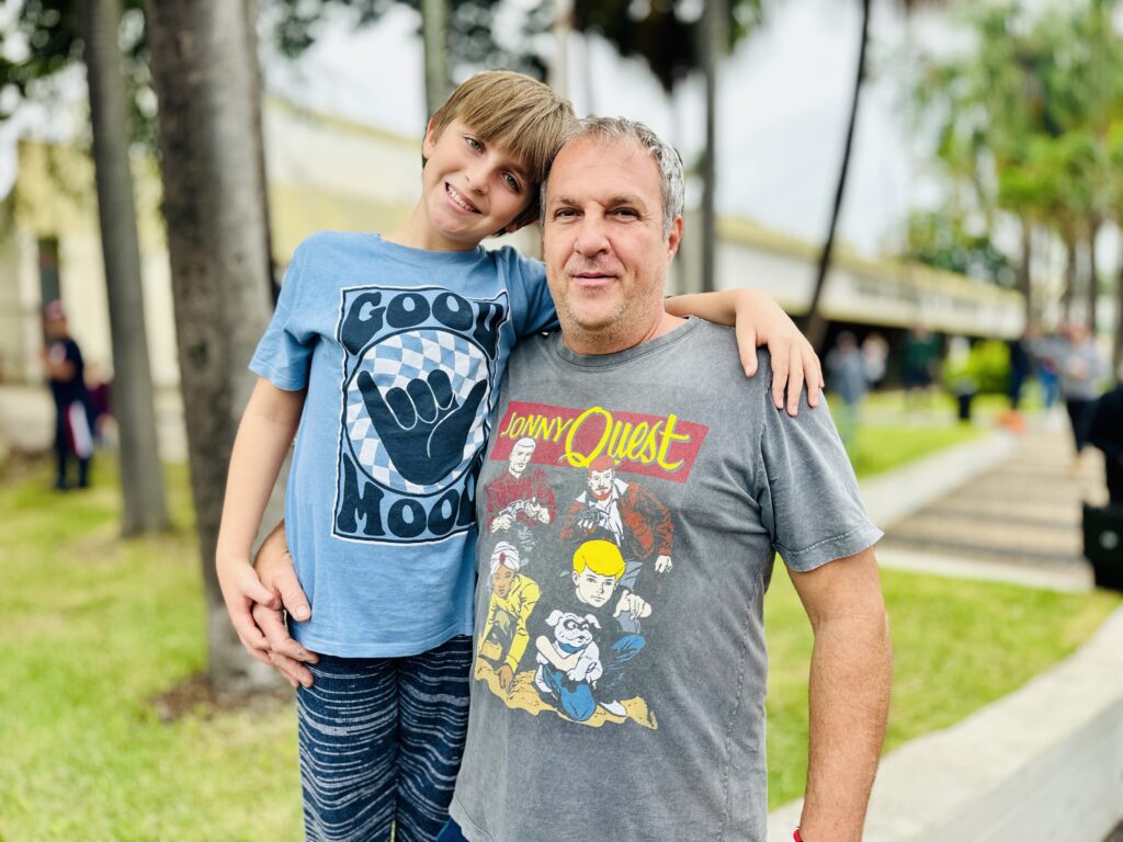 A imagem mostra um homem e um menino juntos ao ar livre, provavelmente em um parque ou área verde. O menino, à esquerda, está sorrindo, tem cabelo castanho claro e veste uma camiseta azul clara com a frase "GOOD MOOD" e um desenho de uma mão fazendo sinal de positivo, além de calças escuras com estampa. Ele apoia o braço sobre o ombro do homem ao lado. O homem, à direita, tem cabelo curto e grisalho, expressão séria ou neutra, e usa uma camiseta cinza com a estampa "JONNY QUEST", que apresenta personagens de desenho animado. Eles estão próximos um do outro, com o braço do homem envolvendo a cintura do menino, transmitindo uma sensação de afeto e proximidade. Ao fundo, há árvores, incluindo palmeiras, e um edifício desfocado, indicando que a foto foi tirada durante o dia, com boa iluminação natural. A imagem tem um foco nítido nos dois, com o fundo desfocado, destacando os sujeitos principais.