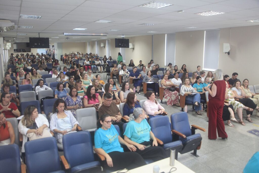 A imagem mostra uma sala ampla, semelhante a um auditório ou sala de conferências, repleta de pessoas sentadas em cadeiras organizadas em fileiras. A maioria dos presentes são mulheres, e todas parecem estar prestando atenção a uma apresentação ou palestra que ocorre na frente da sala. O ambiente é bem iluminado com luz artificial, e as paredes são claras, com janelas grandes que têm persianas. As cadeiras são predominantemente azuis e cinza. No primeiro plano, uma mulher vestindo um macacão vermelho está em pé, segurando um microfone, possivelmente a palestrante ou mediadora do evento. Algumas pessoas estão anotando ou olhando para seus celulares. O clima da imagem sugere um evento educacional, profissional ou um workshop, onde os participantes estão engajados na atividade que está acontecendo. No geral, é uma cena típica de um encontro formal para aprendizado ou troca de informações.