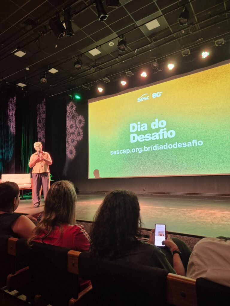 A imagem mostra uma apresentação em um auditório ou teatro. Um palestrante está em pé no palco, segurando um microfone e falando para a plateia. Ele é um homem de cabelos claros, vestido com uma camisa laranja clara e calça escura. Atrás dele, há uma grande tela de projeção com um fundo em degradê que vai do amarelo ao verde, exibindo o texto "Sesc 80 Dia do Desafio" e um endereço de site "sescsp.org.br/diadodesafio". A sala está escura, com algumas luzes no teto direcionadas para o palco. Na plateia, algumas pessoas estão sentadas, observando a apresentação, e uma delas está segurando um celular. O ambiente parece ser profissional e voltado para uma palestra ou evento educativo.