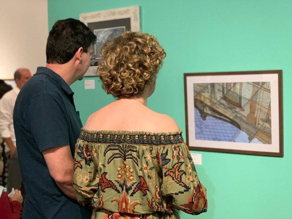 A imagem mostra duas pessoas observando uma obra de arte em uma galeria. À esquerda, um homem de cabelos curtos e escuros, usando óculos e uma camiseta polo azul escura, está de costas para a câmera, olhando para a pintura. Ao centro, uma mulher com cabelos loiros e cacheados, usando uma blusa verde com um padrão colorido e detalhes que lembram folhas de palmeira, também está de costas para a câmera, focada na mesma obra. Eles estão diante de uma parede pintada de um tom vibrante de azul esverdeado. À direita, pendurada na parede, há uma pintura emoldurada, que parece representar uma cena arquitetônica com perspectiva, em tons suaves de marrom, bege e azul. Ao fundo, à esquerda, é possível ver parcialmente outra obra em preto e branco e parte de outra pessoa. A iluminação do local é uniforme, destacando tanto as pessoas quanto as pinturas. A foto foi tirada de trás dos observadores, mostrando a interação deles com a arte.