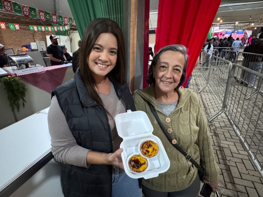 A imagem mostra duas mulheres sorrindo, posando juntas em um ambiente ao ar livre que parece ser uma feira ou festival gastronômico. A mulher à esquerda é mais jovem, tem cabelo castanho longo e está usando uma blusa bege com um colete preto. Ela segura uma embalagem branca de isopor aberta, que contém dois pastéis de nata, uma sobremesa típica portuguesa. A mulher à direita é mais velha, com cabelo grisalho curto, vestindo um suéter verde claro com botões e uma bolsa transversal preta. Ao fundo, há decorações coloridas em verde e vermelho penduradas, além de uma estrutura metálica e uma placa que indica "BOLINHO DE BACALHAU", sugerindo que o evento oferece comidas típicas portuguesas. O chão é de pedras e o ambiente está iluminado artificialmente, indicando que pode ser uma festa noturna ou em um espaço coberto.