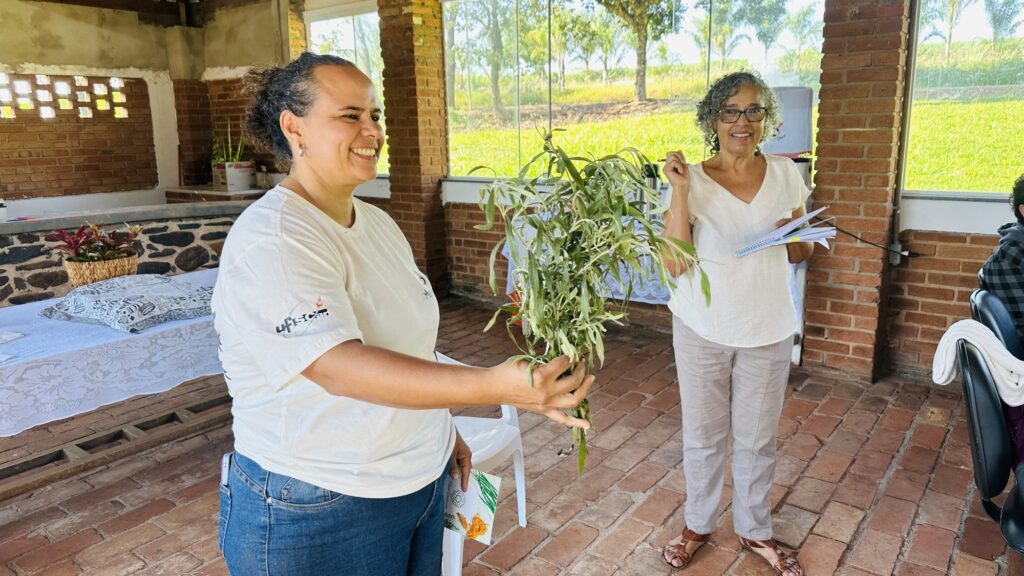 A imagem mostra duas mulheres em um ambiente claro e arejado, com paredes de tijolos aparentes e grandes janelas que dão para uma área verde ao fundo. A mulher em primeiro plano está vestindo uma camiseta branca com o logo "UFSCar" (Universidade Federal de São Carlos) e calça jeans azul. Ela está segurando um maço de ervas verdes na mão direita e um folheto na mão esquerda, sorrindo enquanto olha para o lado direito da imagem. Atrás dela, há outra mulher com cabelos grisalhos e cacheados, usando óculos e uma blusa branca decotada em V, além de calça clara. Ela está segurando um caderno ou documento na mão esquerda e com a mão direita encostada na cabeça, também sorrindo. O ambiente parece ser uma espécie de espaço comunitário ou sala de aula ao ar livre, com piso de cerâmica e uma mesa coberta por uma toalha branca de renda à esquerda da imagem, onde há uma cesta com mais plantas ou ervas. A luz é natural, indicando que a foto foi tirada durante o dia. A atmosfera geral é descontraída e pode estar relacionada a uma atividade educativa, como uma oficina sobre plantas ou cultivo.