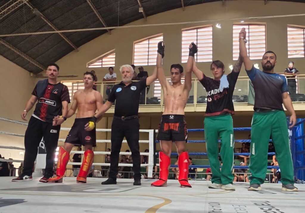 A imagem mostra o momento após uma luta, provavelmente de Muay Thai ou kickboxing, dentro de um ringue de boxe. No centro, dois lutadores estão lado a lado, ambos sem camisa e usando protetores de canela vermelhos. Eles têm os braços levantados por dois árbitros, indicando que a luta acabou e um deles provavelmente é o vencedor. Ao lado dos lutadores, há duas outras pessoas, possivelmente treinadores ou auxiliares, também dentro do ringue. O local parece ser uma arena esportiva com teto alto e algumas janelas, com espectadores assistindo ao redor do ringue. O ringue tem cordas azuis e o piso é branco com algumas marcações. A iluminação é forte, destacando as pessoas no centro do ringue. Os lutadores aparentam estar cansados, mas focados, e os árbitros estão sérios e profissionais. No geral, a cena transmite o momento de anúncio do resultado da luta, com a atmosfera típica de competição esportiva em artes marciais.