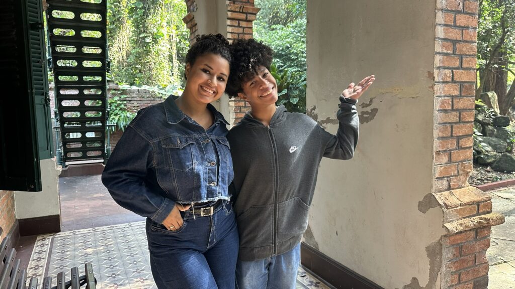 A imagem retrata duas pessoas sorridentes posando lado a lado em um ambiente externo, sob uma varanda com colunas de tijolos aparentes. * A pessoa à esquerda veste uma jaqueta jeans curta com botões metálicos e calça jeans, mantendo uma das mãos no bolso. Seu cabelo está preso em um penteado volumoso. * A pessoa à direita veste um moletom cinza com zíper da marca Nike e calça jeans clara. Ela está com o braço levantado, fazendo um gesto como se estivesse apresentando o ambiente ao redor. * O cenário ao fundo é um espaço aberto com vegetação densa e verde, sugerindo um jardim ou parque. À esquerda, nota-se parte de uma estrutura metálica escada ou grade, e o piso é composto por ladrilhos decorados. A iluminação é natural e suave, indicando um dia ensolarado.