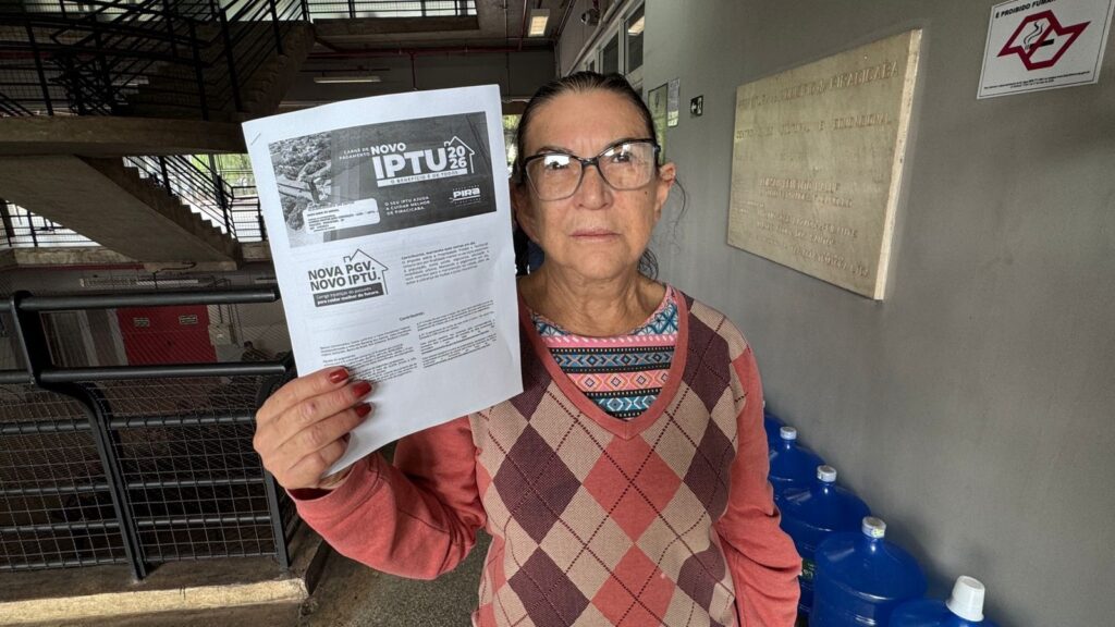 A imagem mostra uma mulher de meia-idade, com cabelos escuros presos, óculos de grau e vestindo um suéter de tricô com estampa de losangos em tons de rosa e marrom. Ela está posicionada centralizada, segurando um documento oficial à frente do corpo com a mão direita. O documento é um carnê de pagamento do IPTU 2026 emitido pela Prefeitura de Piracicaba, contendo o título "NOVA PGV. NOVO IPTU" e informações sobre o pagamento do imposto. O cenário parece ser o interior de um prédio público ou administrativo. Ao fundo, à direita, há uma placa comemorativa fixada na parede, que menciona "Centro Cívico, Cultural e Educacional" e o nome de Piracicaba. Abaixo da placa, observa-se uma fileira de galões de água mineral azuis. À esquerda, nota-se uma estrutura metálica de uma escadaria ou grade. A iluminação é clara e o ambiente sugere um contexto de atendimento ao cidadão.