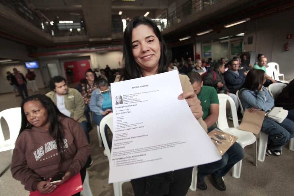 A imagem mostra uma cena bastante comum e cheia de esperança: uma mulher segura um currículo bem à frente, como se estivesse apresentando suas qualificações. Ela parece tranquila e confiante, pronta para uma oportunidade. Ao fundo, dá para ver que ela está em um ambiente que parece um centro de atendimento ou uma agência de empregos, com várias cadeiras ocupadas por outras pessoas que provavelmente também estão aguardando sua vez, cada uma com seus próprios objetivos. O local tem aquele visual de repartição pública ou centro comunitário, com uma atmosfera de expectativa compartilhada.