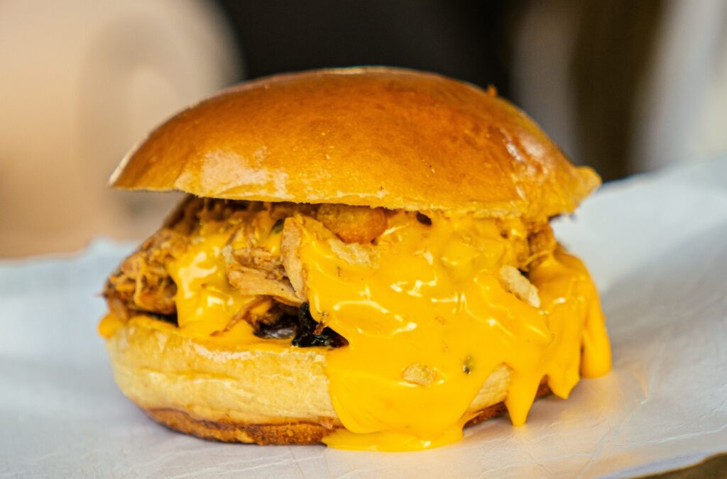 A imagem apresenta um hambúrguer de aparência suculenta, servido sobre uma folha de papel manteiga branco. O pão é do tipo brioche, com uma superfície dourada, brilhante e uniforme. O recheio principal parece ser carne desfiada (possivelmente pulled pork), que está coberta por uma camada generosa de queijo derretido, de cor amarela intensa, que escorre pelas laterais do sanduíche. A iluminação destaca a textura da carne e o brilho do queijo, criando um contraste atraente com o pão, enquanto o fundo permanece desfocado para manter o foco total no alimento.
