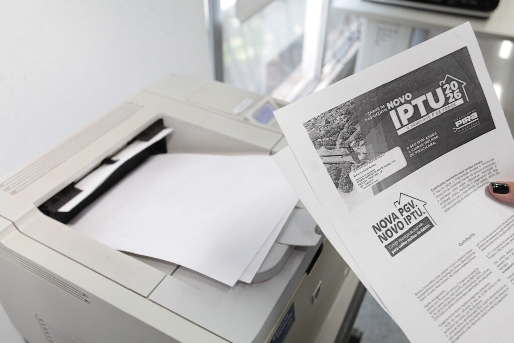 A imagem mostra, em primeiro plano, uma mão segurando uma folha de papel impressa referente ao IPTU 2026 da cidade de Piracicaba. O documento contém informações sobre o pagamento do imposto, incluindo o nome do contribuinte, o endereço e explicações sobre a finalidade da arrecadação, destacando o slogan "O benefício é de todos". Ao lado da mão, observa-se uma impressora laser de mesa com a bandeja de papel aberta, contendo folhas em branco. O ambiente parece ser um escritório ou um espaço de trabalho, com uma iluminação clara e foco nítido no documento que está sendo segurado.