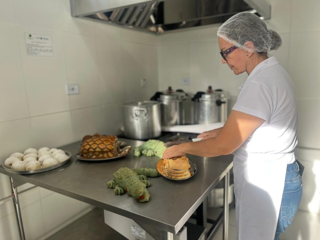 A imagem mostra uma mulher em uma cozinha comercial, cortando um pão em uma mesa de trabalho de aço inoxidável. Ela está usando uma camisa polo branca, avental branco e uma touca descartável para cabelo. Na mesa, além do pão que ela está cortando, há uma bandeja redonda com vários pãezinhos pequenos polvilhados com farinha branca, um pão retangular com cortes em formato de cruz e dois pães verdes com formato que lembra pequenos jacarés. A cozinha possui paredes azulejadas brancas, um exaustor de inox acima da mesa e alguns utensílios grandes, como panelas, ao fundo. A iluminação é clara, provavelmente natural, destacando os detalhes dos pães e da mulher concentrada em seu trabalho.