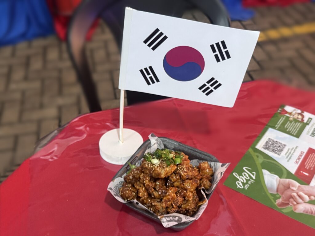 A imagem mostra um prato de frango frito ao estilo coreano, servido em um recipiente quadrado pequeno, forrado com papel personalizado. O frango está coberto por um molho brilhante, provavelmente agridoce ou picante, e decorado com sementes de gergelim e pedaços de cebolinha verde. Ao fundo do prato, há uma pequena bandeira da Coreia do Sul em um suporte branco, indicando a origem ou o estilo do prato. O conjunto está sobre uma toalha vermelha, que destaca as cores do prato. Ao lado direito, há um folheto ou cardápio parcialmente visível, com texto, um código QR e uma ilustração de duas mãos se cumprimentando. O ambiente parece ser ao ar livre ou em um local com piso de pedra, sugerindo um evento ou feira gastronômica. A iluminação é natural, realçando a textura crocante e o brilho do molho no frango.