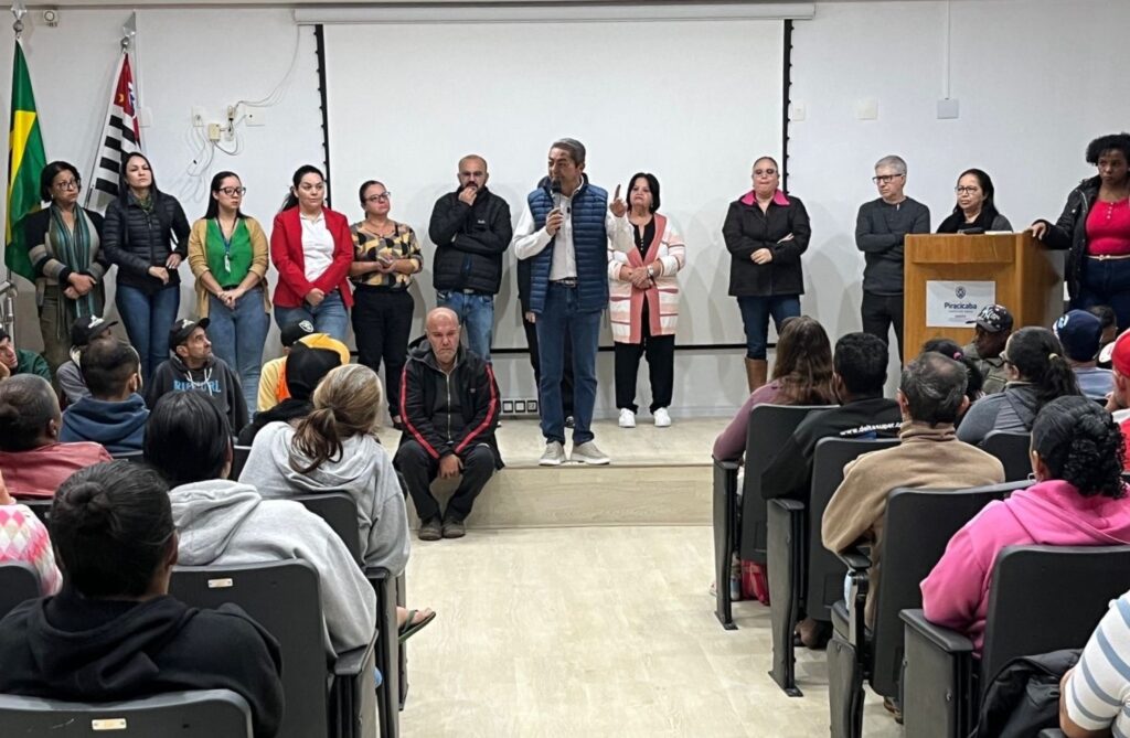 A imagem mostra um ambiente interno, provavelmente uma sala de conferências ou auditório, onde está acontecendo uma apresentação ou reunião formal. No centro, um homem está em pé sobre um pequeno palco, falando ao microfone e gesticulando com a mão esquerda. Ele veste uma camisa branca, colete azul e calça jeans. Atrás dele, em linha, estão cerca de dez pessoas, a maioria mulheres, que parecem estar observando ou aguardando sua vez de falar. À direita da imagem, há um púlpito de madeira com o brasão e o nome "Piracicaba", indicando que o evento está acontecendo nesta cidade. Ao fundo, há uma grande tela branca, provavelmente para projeção de slides ou vídeos. À esquerda, é possível ver duas bandeiras, uma do Brasil e outra do estado de São Paulo. Na frente, estão as cadeiras com o público sentado, assistindo à apresentação. O ambiente é formal, com iluminação clara e organizada, sugerindo um evento institucional ou oficial.