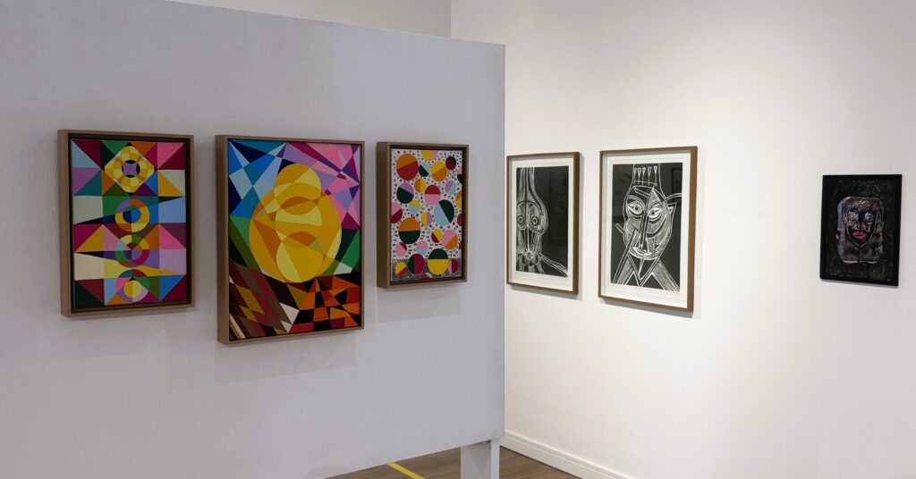 A imagem mostra o interior de uma galeria de arte com várias obras emolduradas expostas nas paredes brancas. À esquerda, há uma divisória onde estão penduradas três obras: a primeira, à esquerda, é uma pintura abstrata geométrica com triângulos e círculos coloridos; a do meio apresenta uma grande forma circular amarela que lembra o sol, sobre um fundo com formas geométricas coloridas; a terceira é uma peça vertical com um padrão de círculos multicoloridos sobre fundo branco. Na parede principal ao fundo, há duas gravuras em preto e branco em estilo xilogravura, retratando rostos estilizados com linhas marcantes e expressivas. À direita, pendurada na parede, está um pequeno retrato escuro, também de um rosto. O ambiente é limpo e minimalista, com paredes brancas e iluminação clara que destaca as obras, criando uma atmosfera profissional e organizada para a exposição. A perspectiva da foto está na altura dos olhos, mostrando claramente a disposição das obras na galeria.