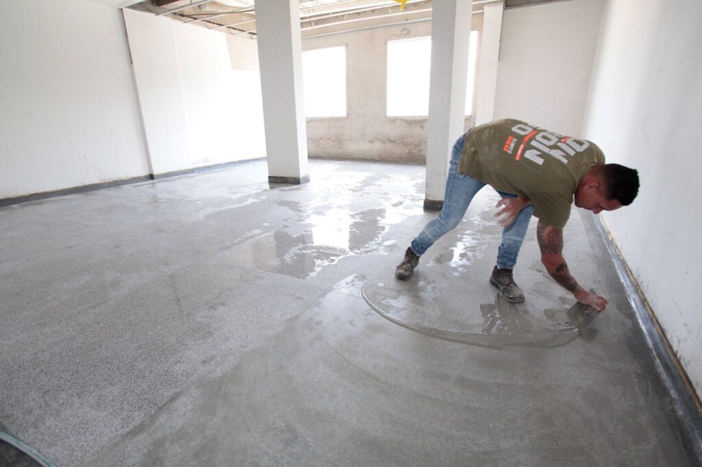A imagem mostra um homem trabalhando na construção de um piso. Ele está aplicando uma camada de argamassa ou um nivelador úmido sobre um piso de concreto em uma sala ampla e ainda em acabamento. As paredes são brancas e sem pintura, e há pilares estruturais visíveis, além de janelas grandes que deixam entrar bastante luz natural. O homem está agachado, usando uma camiseta verde, jeans azuis e botas de trabalho, com tatuagens visíveis nos braços. O piso apresenta áreas já cobertas com o material úmido e outras partes ainda expostas, mostrando o contraste entre o piso bruto e o que está sendo nivelado. A luz do ambiente é clara e uniforme, destacando o trabalho em andamento. No canto superior esquerdo da imagem, há um texto que diz "ALWAYS AHEAD".