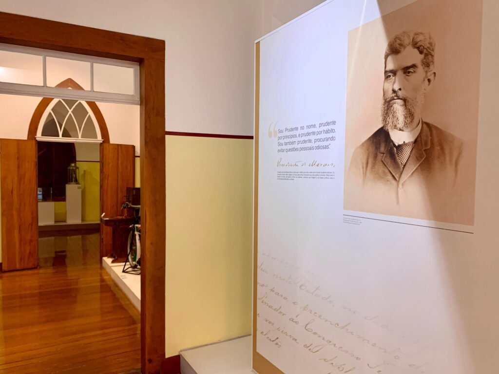 A imagem mostra o interior de um museu ou espaço de exposição histórica. Em primeiro plano, há um grande painel vertical com uma fotografia antiga em sépia de Prudente de Morais, um importante personagem histórico brasileiro. Ao lado da foto, está uma citação dele em português que destaca sua prudência como característica pessoal e de vida. Abaixo da citação, há um texto explicativo que fala sobre sua vida, seus princípios morais, éticos e legais, e sua contribuição para a consolidação da República no Brasil. Também aparece a assinatura de Prudente de Morais e uma reprodução de uma escrita manuscrita antiga. À esquerda, uma porta de madeira aberta permite a visão de uma sala adjacente, com paredes amarelas claras, piso de madeira e uma janela arqueada que deixa entrar luz natural. Dentro dessa sala, é possível ver alguns objetos expostos, como uma bicicleta antiga e uma vitrine com outros itens. A iluminação é suave, criando um ambiente acolhedor e propício para a visitação e aprendizado histórico. A composição da foto utiliza o vão da porta para enquadrar o espaço ao fundo, conferindo profundidade à cena.
