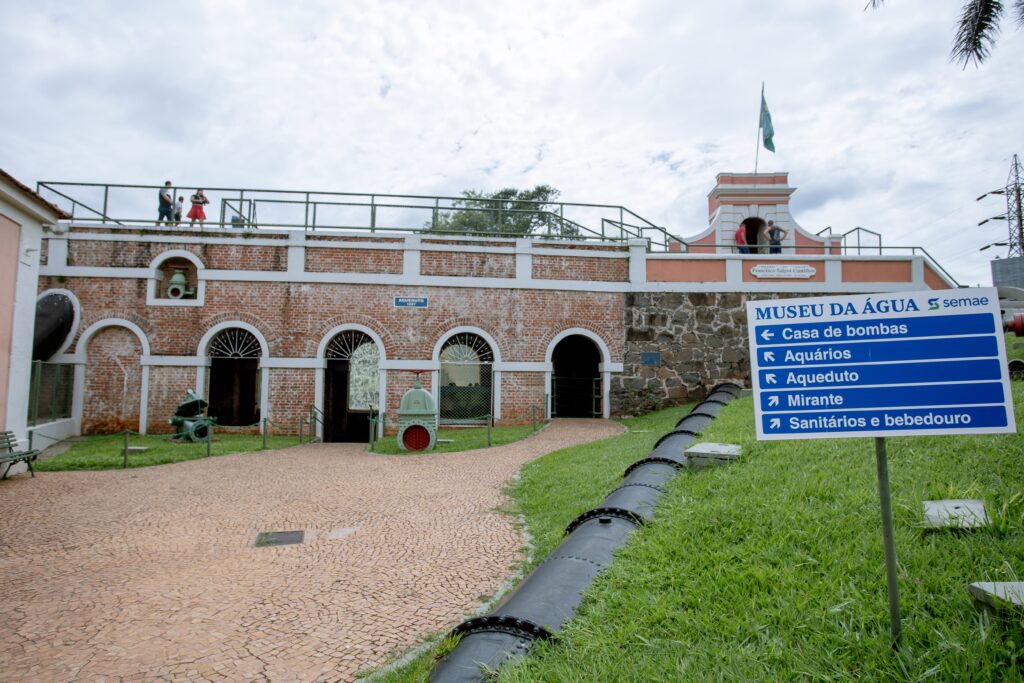 A imagem retrata a fachada do Museu da Água, localizado em Piracicaba, São Paulo. O edifício possui uma arquitetura histórica de tijolos aparentes com arcos, remetendo a uma antiga instalação de tratamento de água. * No centro da imagem, há uma placa informativa azul contendo o nome do local e indicações de direção para áreas como "Casa de bombas", "Aquários", "Aqueduto", "Mirante" e "Sanitários e bebedouro". * À direita da placa, estende-se uma grande tubulação metálica preta, disposta rente ao gramado, que conduz o olhar até a base da estrutura. * Na parte superior da construção, observa-se uma plataforma elevada com guarda-corpo, onde algumas pessoas caminham. No lado direito, há uma pequena torre com uma bandeira hasteada. * O ambiente é iluminado por uma luz natural de dia nublado, com um calçamento de pedras portuguesas em tons terrosos ocupando o primeiro plano à esquerda.