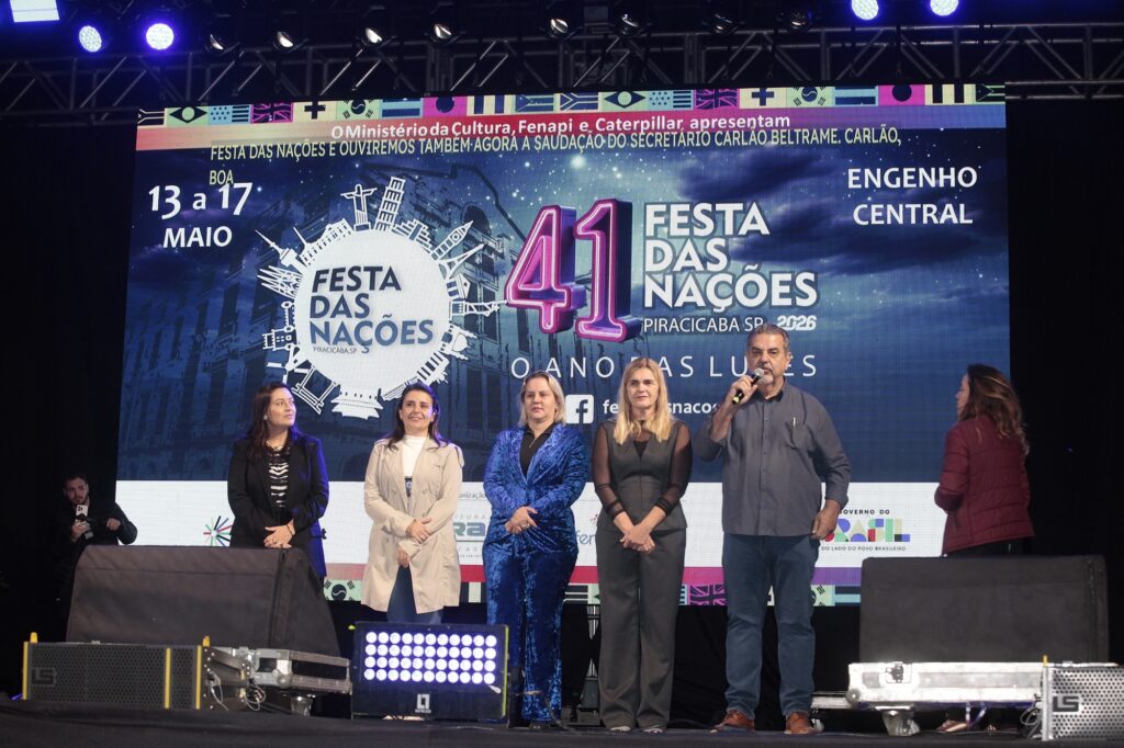 A imagem mostra um evento oficial, possivelmente a cerimônia de abertura da "41ª Festa das Nações" em Piracicaba, São Paulo, que ocorrerá de 13 a 17 de maio no Engenho Central. No palco, estão seis pessoas alinhadas, sendo cinco de frente para o público e uma voltada para elas. Entre os presentes, há quatro mulheres e dois homens, todos vestidos de forma formal ou semiformal. Um dos homens está falando ao microfone, sugerindo que está fazendo uma saudação ou discurso. Ao fundo, há uma grande tela digital com o nome do evento, datas, local e diversas bandeiras nacionais na parte superior, simbolizando a diversidade cultural da festa. Também aparecem logos de patrocinadores e apoiadores, como o Ministério da Cultura e o Governo do Brasil. A iluminação do palco é forte, destacando os participantes e a tela, criando um ambiente de importância e celebração cultural. A cena transmite uma atmosfera de oficialidade e expectativa para o evento que será realizado.