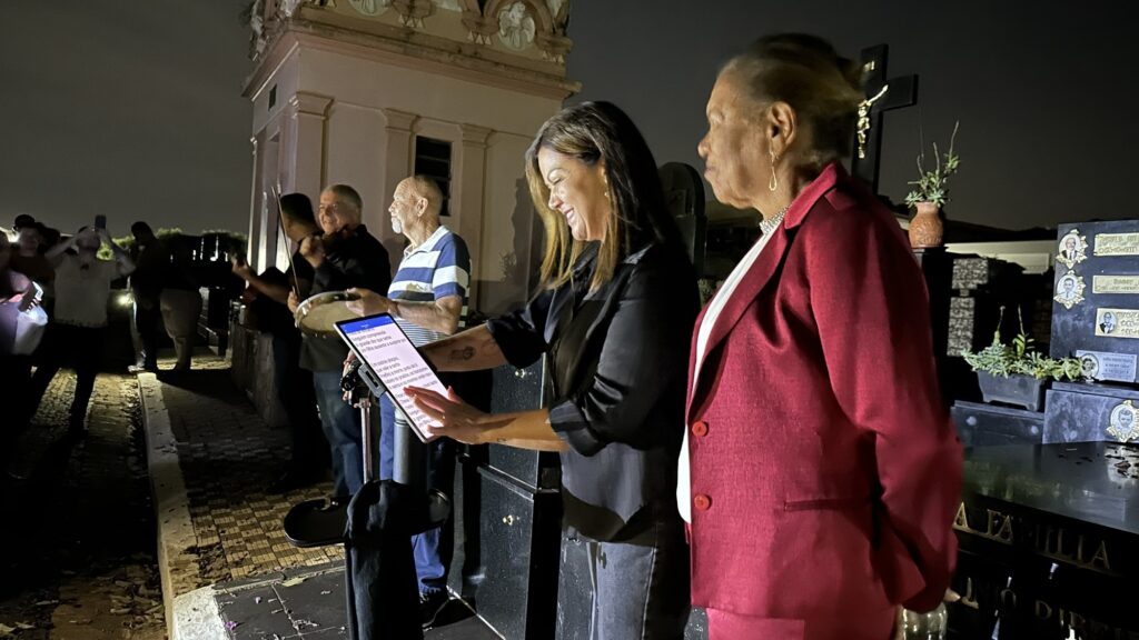 A imagem mostra uma cena noturna em um cemitério, onde um grupo de pessoas está reunido, aparentemente para uma homenagem ou cerimônia musical. No centro da imagem, uma mulher está lendo algo em um tablet apoiado em um pedestal, enquanto outra mulher, vestindo um blazer vermelho, está ao seu lado, possivelmente cantando ou acompanhando a leitura. Ao fundo, alguns homens tocam instrumentos musicais, como um violino e um pandeiro, criando um ambiente de música ao vivo. À direita, há um túmulo de granito preto com várias fotos pequenas de pessoas falecidas fixadas na pedra, sugerindo que a cerimônia é uma homenagem aos mortos. A iluminação artificial destaca as pessoas em primeiro plano, enquanto o fundo permanece mais escuro, dando um tom solene e respeitoso ao encontro. O texto projetado ou exibido na imagem parece ser a letra de uma canção ou poema, expressando sentimentos de saudade e dor pela ausência de um ente querido.