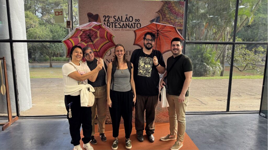 Na imagem aparecem três mulheres e dois homens. No lado esquerdo da foto as duas mulheres seguram uma sombrinha colorida e no outro lado estão dois homens que também seguram um guarda chuva colorido . Eles estão em frente a um prédio de estrutura de vidro e ao fundo aparece o letreiro 22º Salão do Artesanato.
