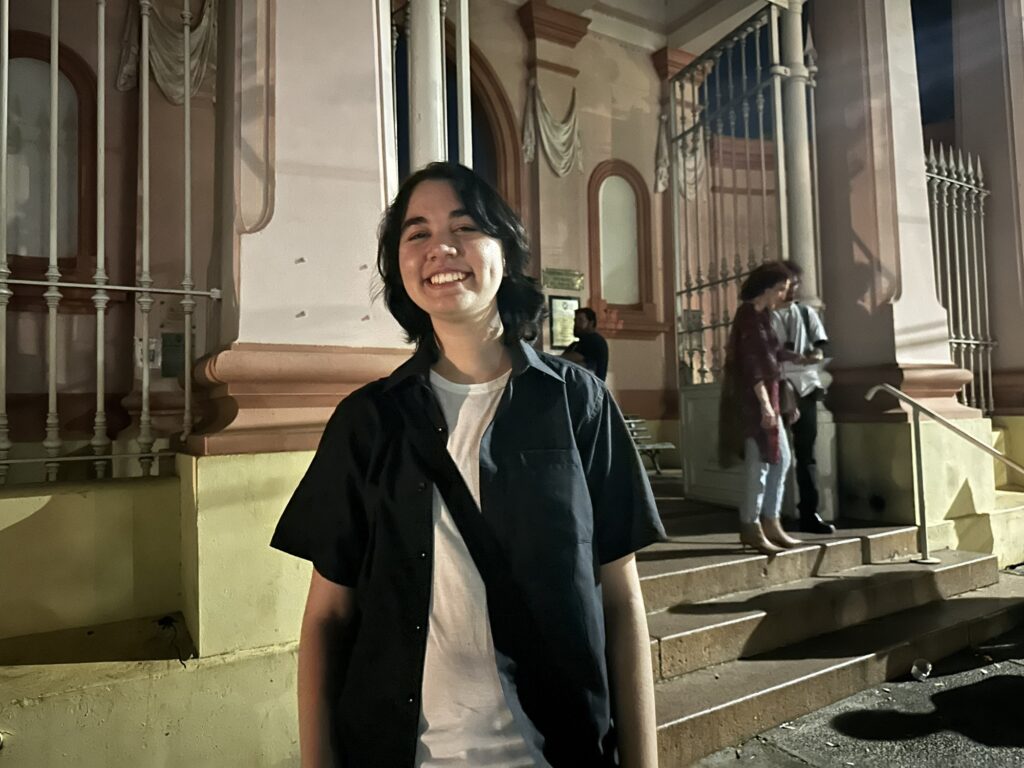 A imagem mostra uma pessoa jovem, de cabelos escuros e lisos na altura dos ombros, sorrindo para a câmera. Ela veste uma camiseta branca por baixo de uma camisa preta de mangas curtas, aberta na frente. A pessoa está em um ambiente externo, à noite, diante de um edifício antigo com detalhes arquitetônicos clássicos, como janelas altas com grades brancas e uma fachada clara com ornamentos. Ao fundo, à direita, há duas outras pessoas próximas à entrada do prédio, que possui degraus de pedra e um portão de metal. A iluminação é artificial, criando um ambiente acolhedor e destacando os contornos da cena.