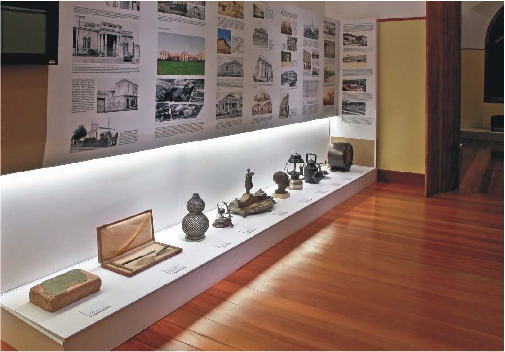 A imagem mostra uma exposição em um museu ou espaço cultural, apresentando uma longa prateleira branca iluminada por baixo, onde estão expostos diversos objetos históricos. Entre eles, há uma peça que parece ser um tijolo antigo, uma caixa forrada com veludo contendo uma ferramenta pequena, um recipiente decorativo em formato de cabaça, uma escultura metálica, um tinteiro ornamentado, uma pequena estátua de busto, uma lanterna antiga e um aparelho que lembra um rádio ou alto-falante antigo. Ao fundo, há um painel alto e branco com várias fotografias em preto e branco e textos explicativos, organizados em colunas, que parecem contar a história local ou mostrar a evolução arquitetônica da região. O piso é de madeira clara e polida, conferindo um ambiente acolhedor e bem cuidado. A iluminação é focada nos objetos expostos, destacando-os contra o fundo branco. À direita, uma porta aberta leva a outro ambiente do museu. A perspectiva é ampla e levemente elevada, proporcionando uma visão geral da exposição.