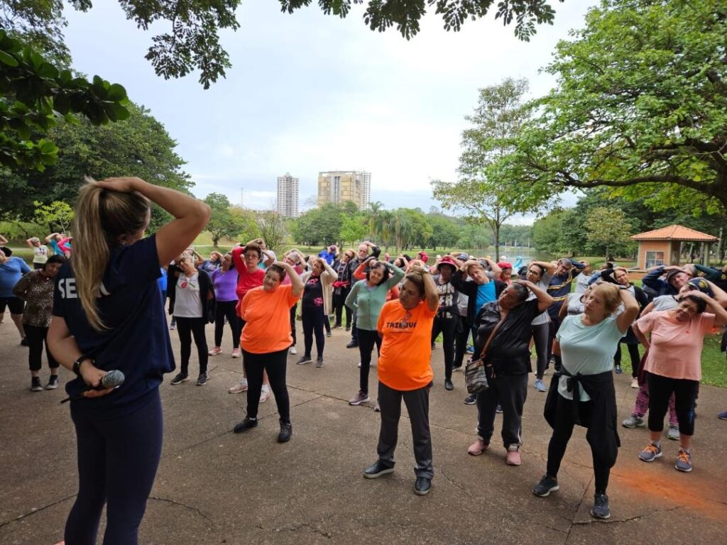 A imagem retrata uma aula de ginástica ao ar livre, em um parque arborizado, composta majoritariamente por um grupo de idosos. * Em primeiro plano, vemos de costas uma instrutora vestindo uma camiseta azul escura com a inscrição "SESC", segurando um microfone e demonstrando um exercício de alongamento com as mãos atrás da cabeça. * O grupo de alunos, posicionado à frente da instrutora, segue o movimento, também mantendo as mãos atrás da cabeça. Eles vestem roupas esportivas variadas, incluindo camisetas de cores vibrantes como laranja e rosa. * O cenário é um ambiente amplo e pavimentado, cercado por árvores, com uma área verde e um lago ao fundo. É possível observar prédios distantes na linha do horizonte, sob um céu nublado. A atmosfera é de uma atividade física comunitária e orientada para a saúde e bem-estar.