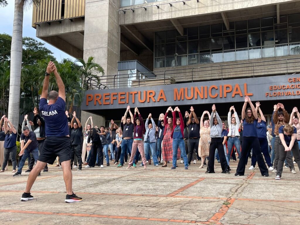 A imagem mostra um grupo grande de pessoas participando de uma atividade física coletiva ao ar livre, provavelmente uma sessão de ginástica laboral ou exercício em grupo promovido pela prefeitura. O local é uma praça em frente a um edifício público, identificado como "Prefeitura Municipal" por letras grandes e laranjas na fachada do prédio. As pessoas estão organizadas em fileiras e parecem estar seguindo um instrutor que está de costas para a câmera, usando uma camiseta azul com a palavra "SELAM" nas costas. Todos estão com os braços levantados, realizando um alongamento ou exercício de aquecimento. O ambiente é urbano, com um prédio grande de arquitetura brutalista ao fundo e algumas palmeiras ao lado esquerdo da imagem. O clima parece ameno, possivelmente em um dia nublado ou com luz suave. A atividade sugere uma iniciativa de promoção da saúde e bem-estar dos servidores municipais ou da comunidade local.