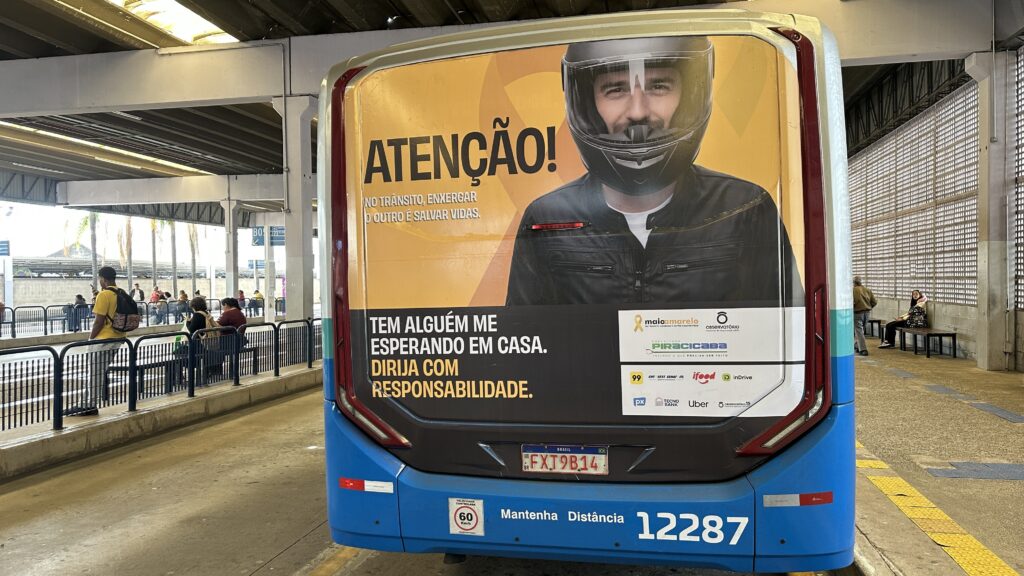 A imagem mostra a parte traseira de um ônibus urbano estacionado em um terminal de transporte. No fundo do ônibus há um grande anúncio publicitário voltado para a conscientização sobre segurança no trânsito. No anúncio, há a foto de um homem usando capacete de motociclista, olhando diretamente para frente. Ao seu lado, está escrito em letras grandes e amarelas: "ATENÇÃO! NO TRÂNSITO, ENXERGAR O OUTRO É SALVAR VIDAS." Logo abaixo, há outra frase em branco: "TEM ALGUÉM ME ESPERANDO EM CASA. DIRIJA COM RESPONSABILIDADE." O anúncio também exibe vários logos de apoiadores e patrocinadores da campanha, como "Maio Amarelo", "Prefeitura de Piracicaba", "99", "iFood", "inDrive", "Uber", entre outros. Na parte inferior do ônibus, há a placa do veículo (FXT9B14, indicando que é do Brasil), uma placa que indica a velocidade máxima controlada de 60 km/h e a frase "Mantenha Distância". O número do ônibus, 12287, está visível próximo à parte inferior. O terminal onde o ônibus está estacionado tem um piso de concreto, cobertura metálica e algumas pessoas aguardando em bancos ao fundo. O ambiente é aberto e iluminado naturalmente.