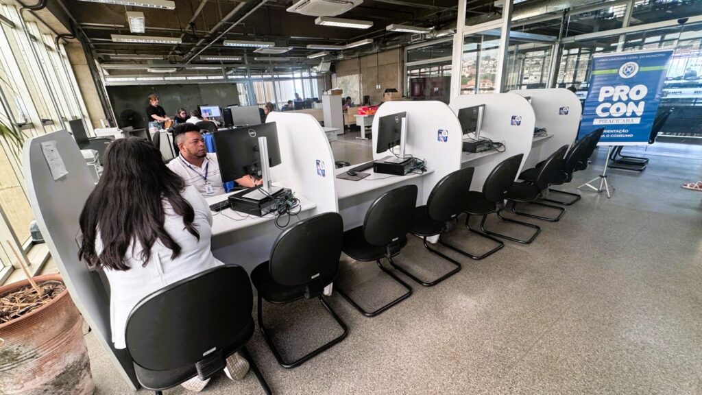 A imagem mostra o interior de um escritório público, provavelmente um órgão de defesa do consumidor, dado o banner visível com os dizeres "PRO CON PIRACICABA ORIENTAÇÃO AO CONSUMO". No centro da imagem, há uma fileira de estações de trabalho com computadores Dell, separadas por divisórias brancas. Uma mulher está sentada em uma das estações, de costas para a câmera, enquanto um homem, provavelmente um funcionário, está sentado em outra estação, olhando para a tela do computador. O ambiente é moderno e funcional, com piso de concreto, cadeiras de escritório pretas e iluminação fluorescente no teto, que também exibe dutos de ar condicionado aparentes. Ao fundo, outros funcionários trabalham em suas estações, indicando um ambiente ativo. A iluminação natural entra pelas janelas, complementando a iluminação interna. O espaço transmite organização e é voltado para o atendimento ao público, especialmente para orientação e suporte ao consumidor.