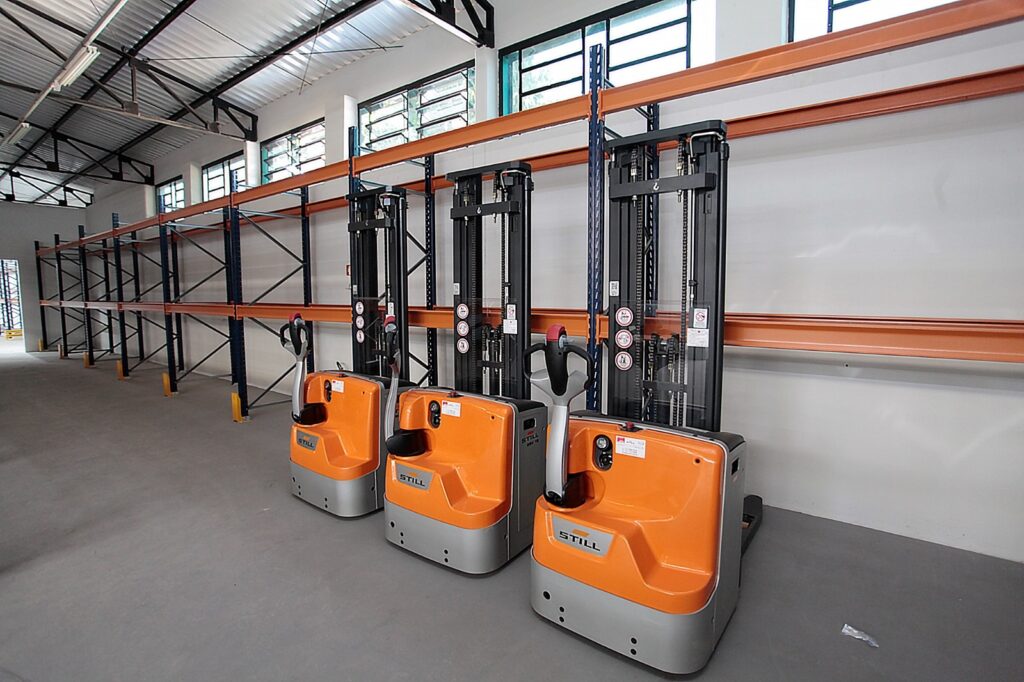 A imagem mostra o interior de um armazém industrial com três empilhadeiras elétricas da marca STILL alinhadas lado a lado. As empilhadeiras são predominantemente laranja com detalhes cinza e possuem uma estrutura vertical preta para elevação. O ambiente é amplo, com piso de concreto cinza, paredes brancas e uma estrutura metálica no teto. Ao fundo, há estantes industriais metálicas de armazenamento, com vigas laranja e colunas azul-escuras. A iluminação parece ser natural, vinda de janelas altas na parede. O espaço está organizado, limpo e vazio de pessoas, transmitindo uma sensação de ordem e eficiência no ambiente de trabalho.