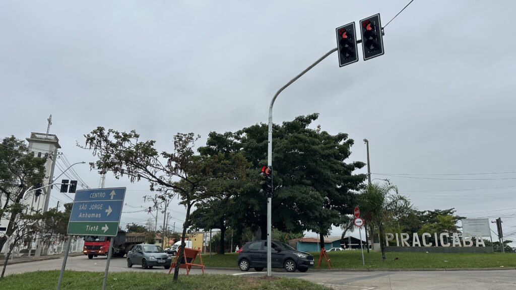 A imagem mostra um cruzamento de ruas na cidade de Piracicaba, com um céu nublado e carregado. No centro, há um semáforo com luz vermelha acesa, indicando que os veículos devem parar. À esquerda, há uma placa azul de sinalização indicando direções para o Centro, São Jorge, Anhumas e Tietê. Na via, observa-se um caminhão vermelho e um carro cinza à esquerda, além de um carro preto parado no semáforo ao centro. À direita, há um grande letreiro de pedra ou concreto com o nome "PIRACICABA", colocado em um gramado, próximo a uma placa de "PARE". Também é possível ver uma placa com um aviso da prefeitura que proíbe o comércio ambulante sem licença. Ao fundo, árvores verdes e postes com fios elétricos completam a paisagem urbana. O ambiente transmite uma sensação de cidade tranquila em um dia nublado.