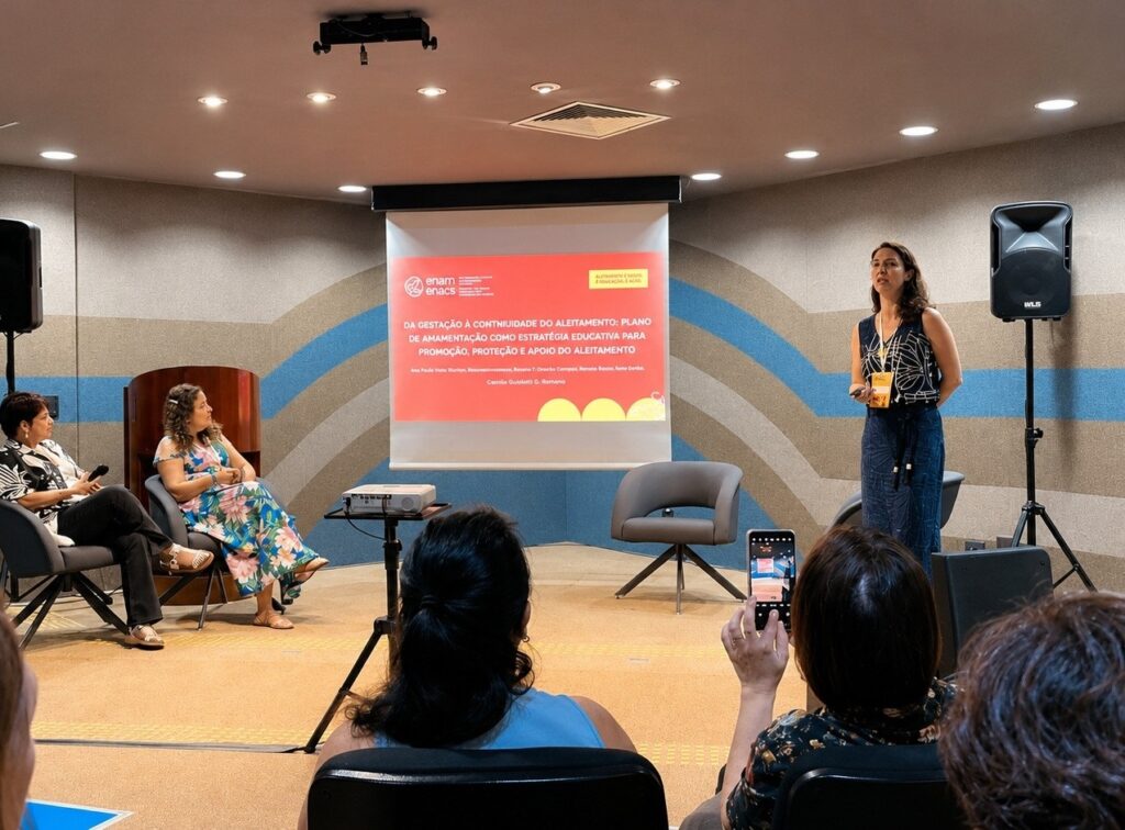 A imagem mostra uma apresentação em um ambiente fechado, possivelmente uma sala de conferências ou aula. À direita, uma mulher está em pé, falando e gesticulando, vestindo uma blusa preta e uma saia escura, com um crachá amarelo pendurado no pescoço. À esquerda, duas mulheres estão sentadas, olhando para a tela de projeção. Uma delas usa uma blusa preta e branca, e a outra, um vestido floral. Na tela de projeção, há um texto em português que fala sobre um plano de amamentação como estratégia educativa para promoção, proteção e apoio do aleitamento, indicando que o tema da apresentação é relacionado à saúde materno-infantil. Também aparecem alguns nomes, possivelmente dos autores ou palestrantes. A sala tem uma decoração com padrões geométricos nas paredes e dois alto-falantes grandes estão posicionados nas laterais. Algumas pessoas da audiência aparecem de costas, e uma delas está segurando um celular para registrar o momento. A iluminação é artificial, típica de ambientes internos para apresentações.