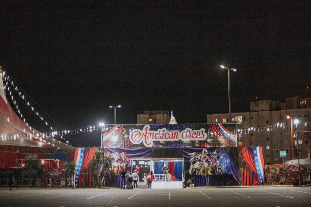 A imagem apresenta a entrada do "American Circus" durante a noite, em um ambiente externo que parece ser um estacionamento vazio. * O elemento central é uma grande fachada iluminada com o nome "American Circus" em letras estilizadas. Abaixo do letreiro, há painéis decorativos que exibem imagens de artistas circenses, incluindo dançarinas e motociclistas em um globo da morte. * À esquerda, é possível ver parte da lona do circo, que é vermelha e está adornada com luzes em série que se estendem para cima. * Em frente à entrada, algumas pessoas estão reunidas, sugerindo que o local está aberto ou prestes a receber público. O cenário é iluminado por postes de luz e luzes decorativas suspensas, criando um contraste com o céu noturno escuro. * Ao fundo, à direita, avista-se um prédio residencial, indicando que o circo está instalado em uma área urbana. A cena transmite uma atmosfera de entretenimento noturno clássico.