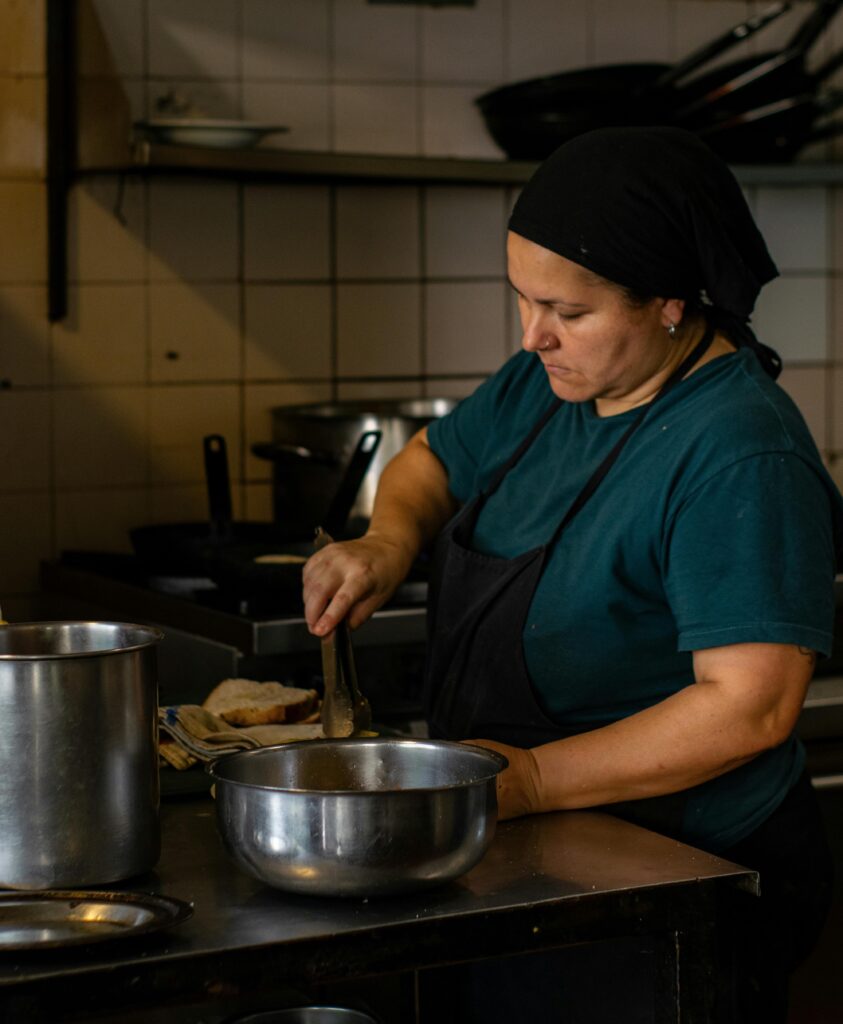 A imagem retrata uma cozinheira em seu ambiente de trabalho, focada na preparação de alimentos. * Ela está posicionada atrás de uma bancada de aço inoxidável, vestindo uma camiseta azul-escura e um avental preto, com um lenço ou bandana preta cobrindo parte da cabeça. * Com o auxílio de um pegador metálico, a profissional manuseia ingredientes dentro de uma tigela de inox posicionada à frente. * Ao fundo, é possível observar uma cozinha profissional com azulejos brancos nas paredes, panelas sobre o fogão e utensílios dispostos em prateleiras, criando um ambiente de trabalho estruturado e funcional.