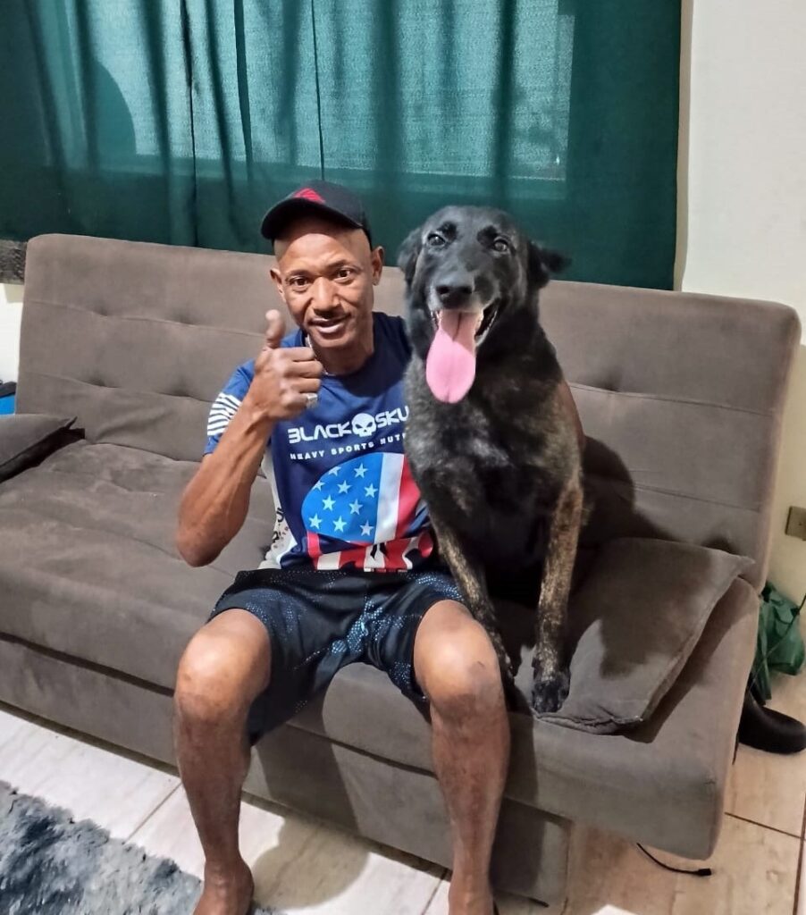 A imagem mostra um homem sentado em um sofá marrom ao lado de um cachorro grande. O homem está usando um boné preto, uma camiseta azul escura com a estampa "BLACK SKULL HEAVY SPORTS NUTRITION" e shorts pretos. Ele sorri e faz um sinal de positivo com a mão direita. O cachorro, que é de porte grande e tem pelagem escura com manchas, está sentado ao lado do homem, com a língua para fora, parecendo calmo e contente. Ao fundo, há uma cortina verde escura. A cena transmite uma sensação de conforto e amizade entre o homem e o animal.