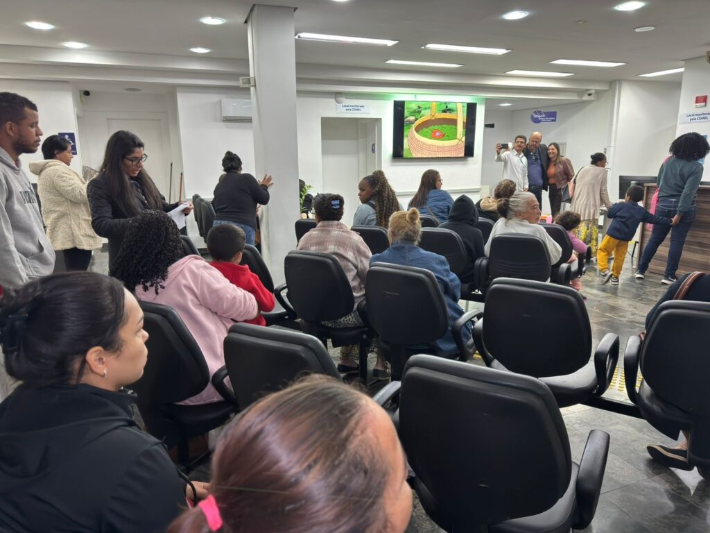 A imagem mostra uma sala de espera ou recepção, possivelmente em uma clínica ou repartição pública, com várias pessoas sentadas em cadeiras pretas dispostas em fileiras. * No centro da sala, há uma tela grande exibindo uma animação colorida, que parece ser um desenho animado com uma cesta sobre grama. * Várias pessoas estão sentadas de costas ou de perfil, aguardando atendimento. No lado direito, um grupo de pessoas, incluindo um homem de camisa branca, está tirando uma foto ou selfie, enquanto uma criança caminha pelo espaço. * O ambiente possui iluminação embutida no teto, paredes brancas e um piso de tom escuro. Há placas indicativas nas paredes, como "Local monitorado pela CEMEL" e "Espaço de Leitura". * A atmosfera é de uma sala de espera comum, com um misto de pessoas sentadas, algumas conversando e outras observando o ambiente ou a tela.