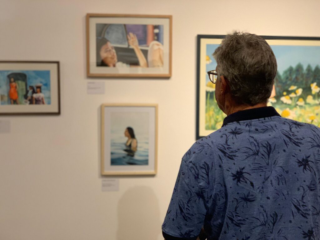 A imagem mostra um homem visto de costas, observando várias pinturas emolduradas expostas em uma galeria de arte. Ele tem cabelos grisalhos, usa óculos e uma camisa azul com estampa de folhas escuras. À sua esquerda, na parede branca, há três quadros: o superior mostra uma pessoa dentro de um veículo com a mão levantada; o do meio retrata uma pessoa em pé dentro da água; e o inferior apresenta figuras em um ambiente ao ar livre, com uma pessoa vestindo laranja em destaque. À direita do homem, há uma pintura maior, parcialmente visível, que mostra uma paisagem com flores amarelas e árvores verdes. A iluminação do local é suave, típica de um ambiente interno de galeria. Pequenas etiquetas brancas estão fixadas abaixo das obras, provavelmente com informações sobre cada pintura.