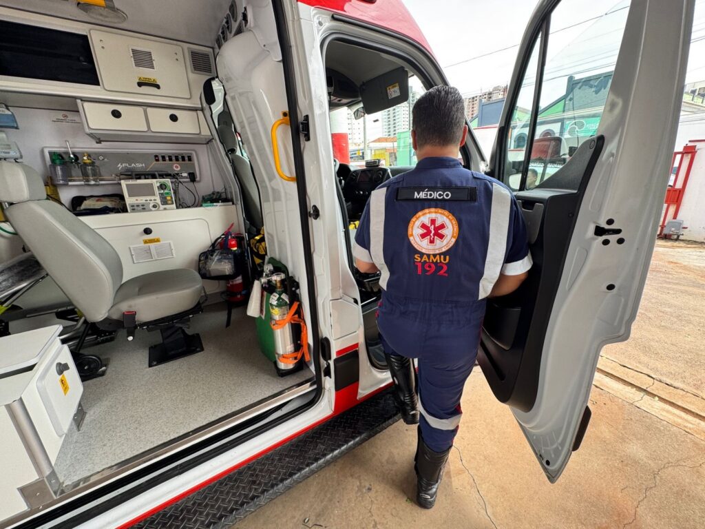 A imagem mostra a parte interna e externa de uma ambulância, com um profissional de saúde vestido com um macacão azul escuro, listrado em branco e vermelho nas mangas e ombros, de costas para a câmera, entrando ou saindo do veículo. No macacão, destaca-se um emblema circular com a Estrela da Vida em vermelho e a inscrição "SAMU 192", além da palavra "MÉDICO" em um patch preto acima do emblema. A ambulância está aberta, revelando um interior bem equipado com equipamentos médicos, incluindo um monitor/desfibrilador identificado pela palavra "FLASH", cilindros de oxigênio, extintores e vários compartimentos de armazenamento. As paredes e o teto internos são de cor branca, o chão é cinza com um padrão pontilhado, e há um assento médico cinza instalado. O ambiente ao redor sugere ser uma área urbana, com vegetação e prédios ao fundo, sob luz natural do dia. A imagem transmite a ideia de prontidão e ação dos serviços de emergência médica, especificamente do SAMU (Serviço de Atendimento Móvel de Urgência) no Brasil.